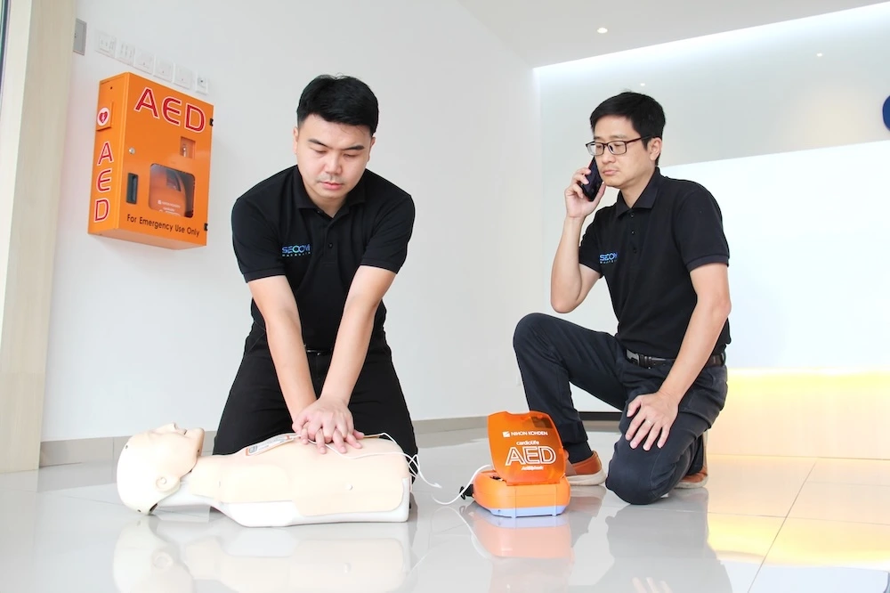 SECOM Malaysia staff performing CPR during AED training while a colleague calls emergency services