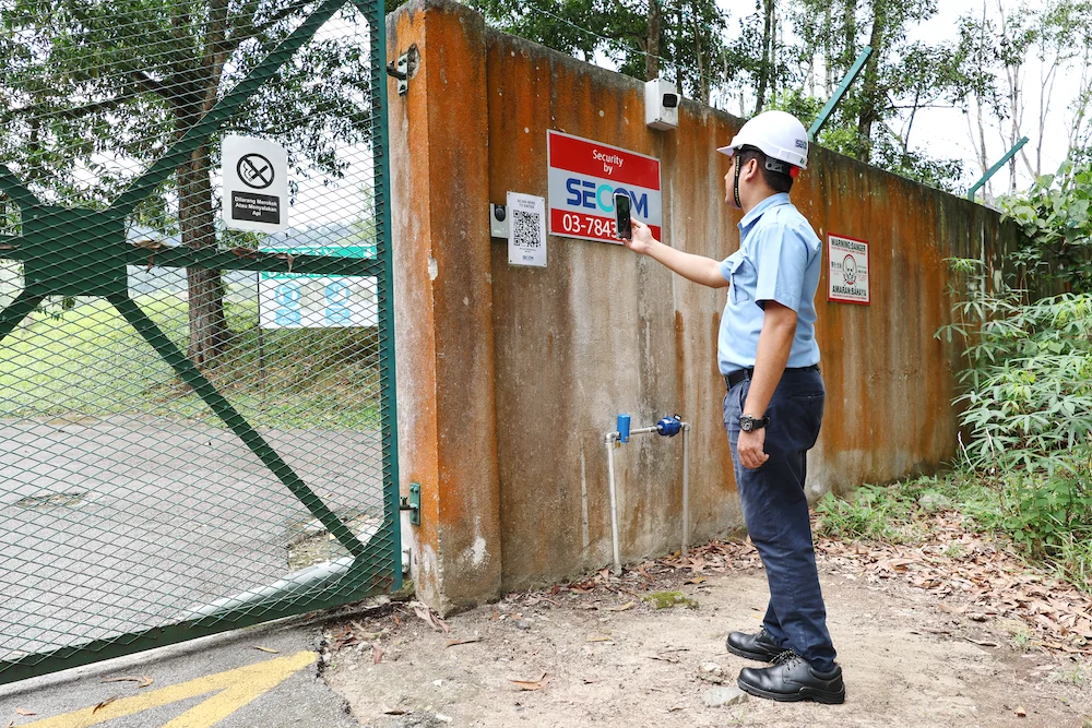 SECOM Malaysia security officer inspecting a factory perimeter security system with secured fencing, access gate and surveillance equipment.