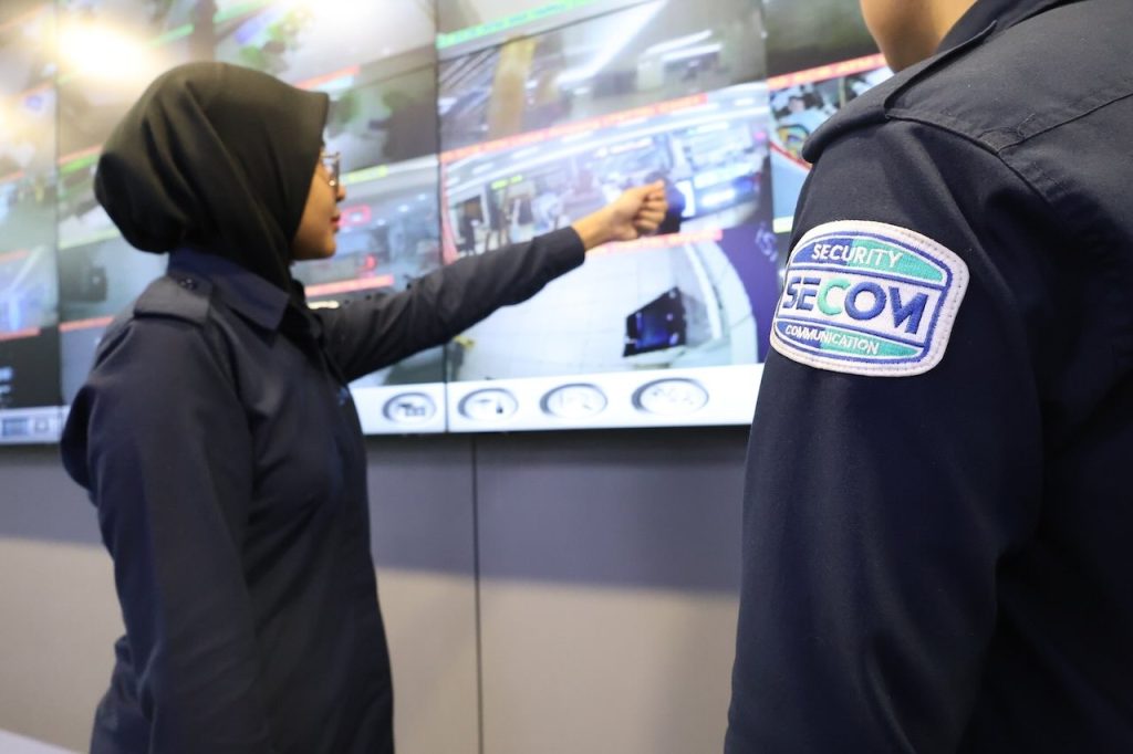 A SECOM security officer holds a smartphone showing live CCTV surveillance footage on its screen, illustrating the convenience and flexibility of remote monitoring systems for security management beyond traditional control room settings.