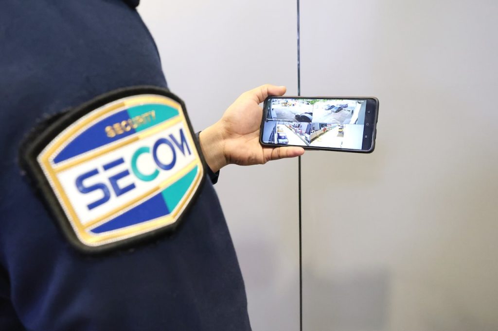 Security personnel in SECOM uniforms stand in front of a large video wall displaying multiple CCTV camera feeds, as one team member points to an incident on the screen. The scene highlights collaborative monitoring, real-time response, and the integration of advanced remote monitoring solutions in a professional facility.