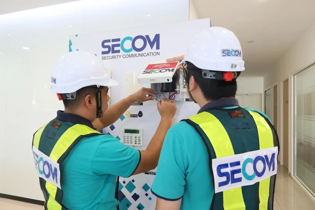 Two SECOM Malaysia technicians installing and configuring an alarm control panel in a building security system setup.