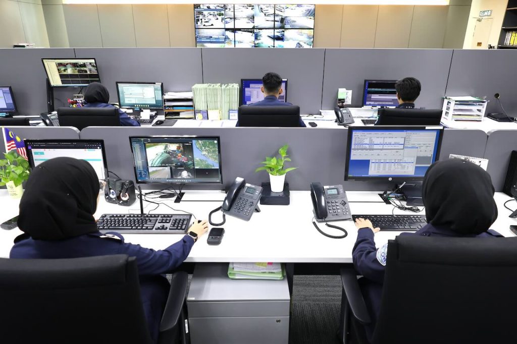 Row of SECOM security operators monitoring a large video wall of CCTV cameras in a central monitoring system control room.
